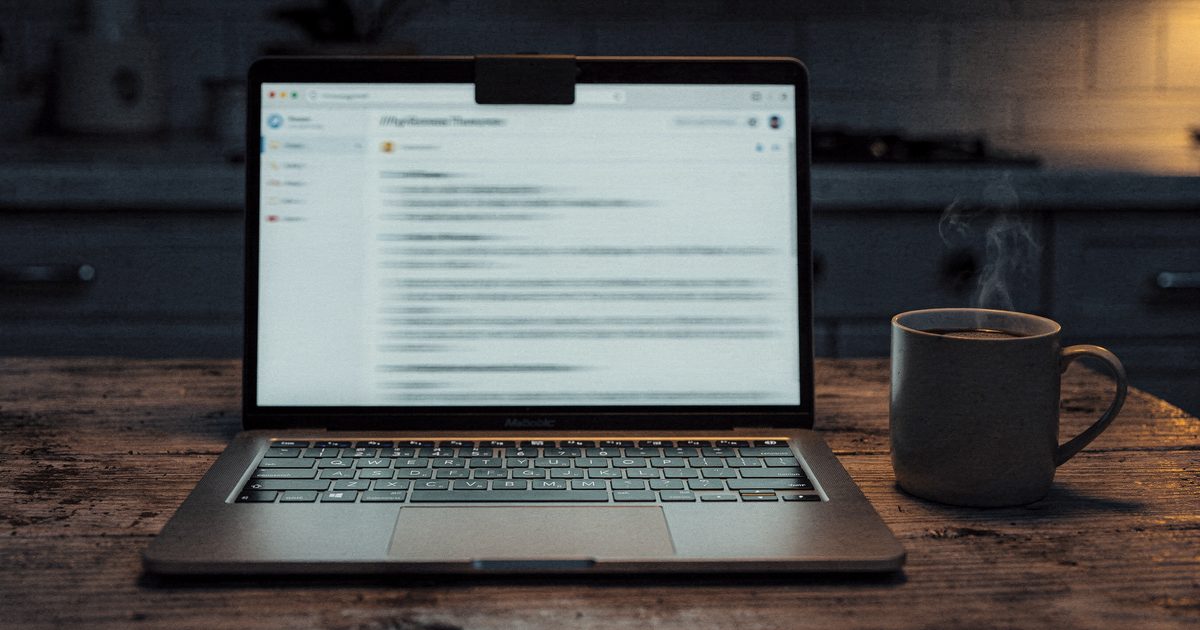 A laptop on a kitchen table at night showing an open email with a blurred threatening message, a mug of coffee beside it and a closed webcam cover on the laptop lid