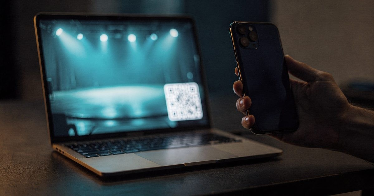 A laptop on a dark desk at night playing a blurred livestream with a glowing QR code in the corner and a large viewer counter, a phone beside it held up as if to scan the code