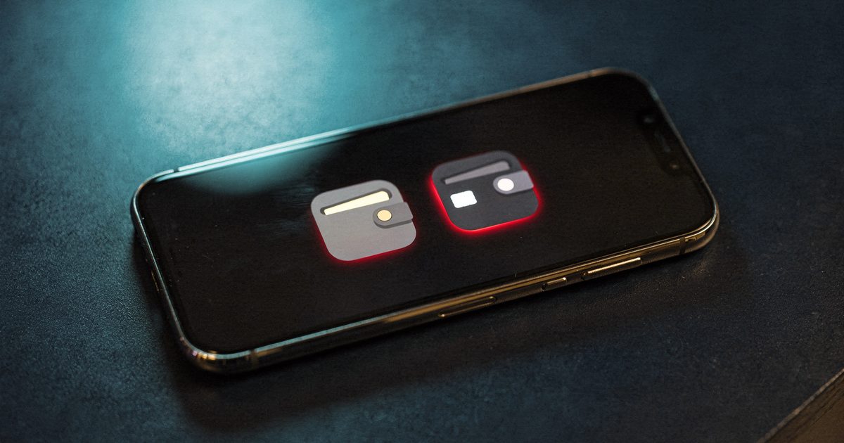 A smartphone on a dark desk showing two nearly identical wallet app icons side by side, one with a faint red glow, under a cool teal desk light