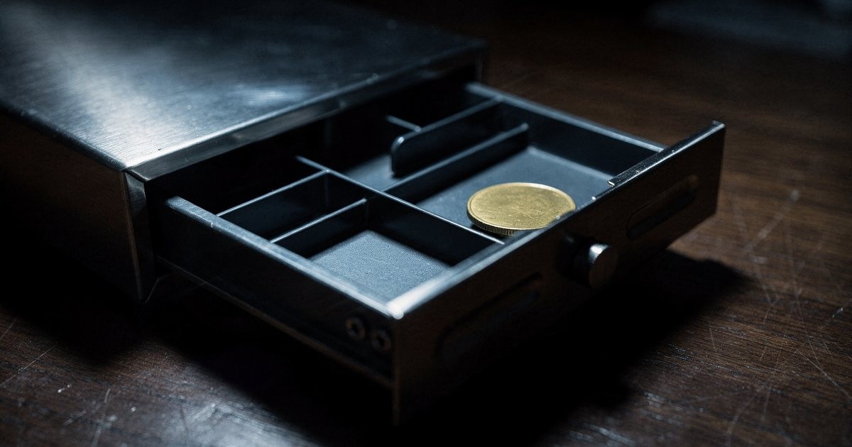 A dark steel cash drawer pulled open on a wooden desk, a single gold coin left inside, lit from above