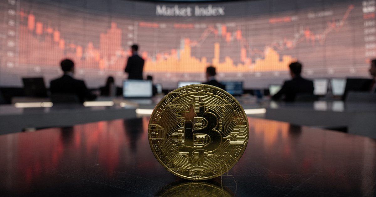 A gold bitcoin on a desk in front of trading screens showing red falling charts