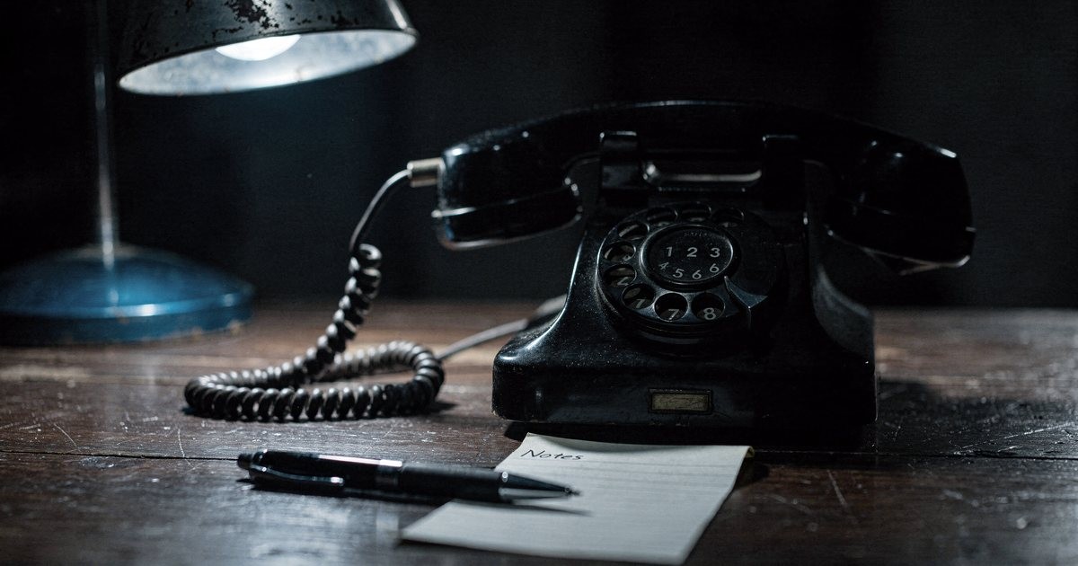 An old black rotary telephone on a dark wooden desk under a cold blue lamp, with a pen and a notepad in front of it
