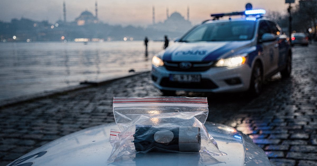 A quiet Istanbul street at dawn with a police car's blue lights and an evidence bag holding a hardware wallet