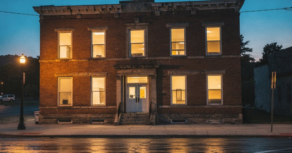 A small-town bank front on a quiet main street at dusk, warm light in the window
