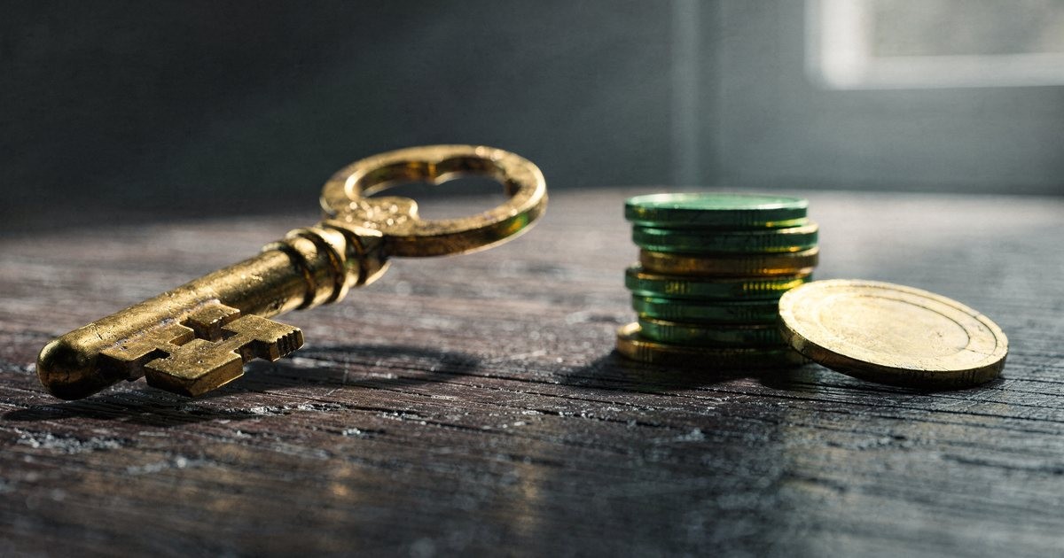 A heavy old brass key on a dark wooden table beside a small stack of plain coins and one large gold coin