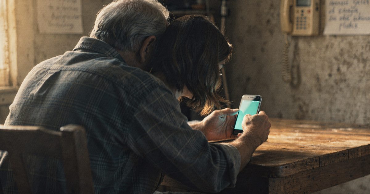 An older man and his adult daughter at a kitchen table looking at a smartphone together, a handwritten note taped to a wall phone behind them, warm afternoon light