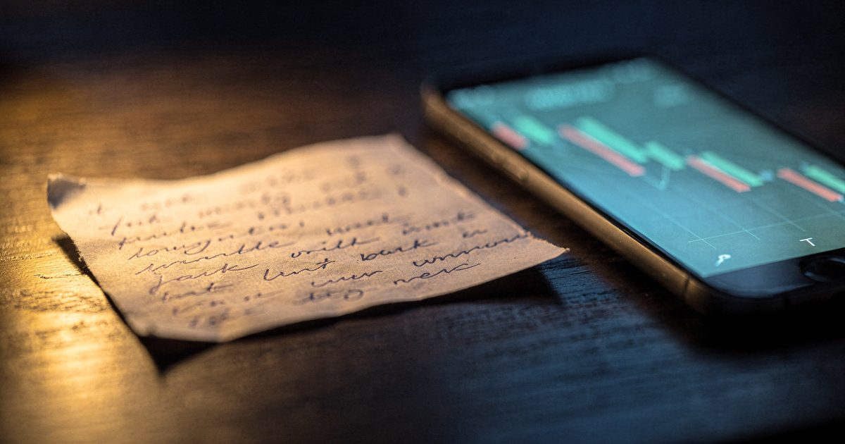 A hand holding a handwritten seed phrase card under a desk lamp while a phone beside it shows a wallet balance draining to zero